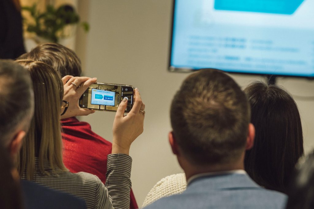 Sieviete koferenču zālē fotografē pasākum anorises laikā prezentāciju uz lielā ekrāna/